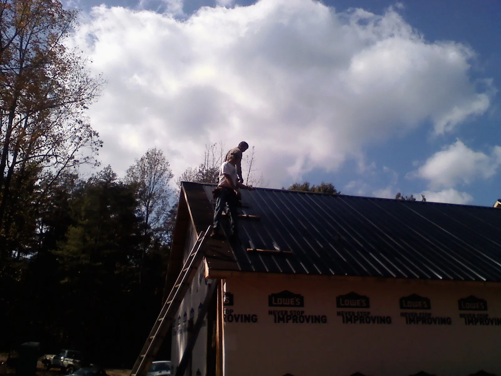 Roofer fastening dark metal panels high against a cloudy sky, from a Roof Inspection job in Savage